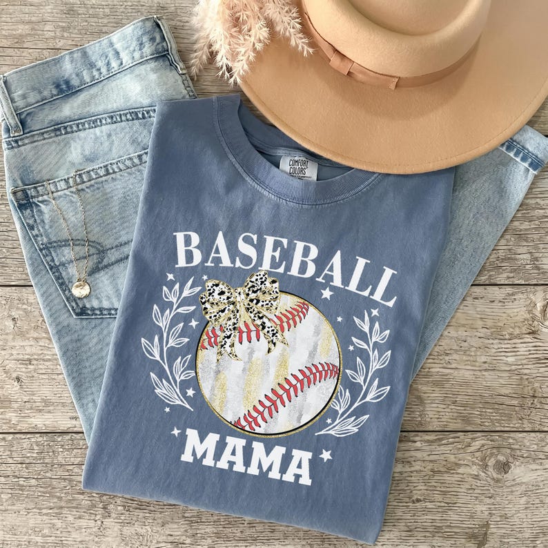 May include: A blue t-shirt featuring the white text BASEBALL MAMA, a graphic of a baseball with red stitching, a leopard print bow, and white leaf accents. The shirt is displayed with light blue denim jeans, a tan hat, and a gold necklace.
