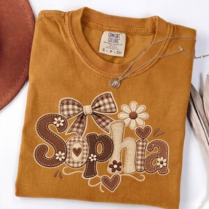 Puede incluir: Una camiseta de color amarillo mostaza con el nombre Sophia bordado en tonos marrón y beige. El diseño incluye un lazo con estampado de cuadros vichy, pequeñas margaritas y formas de corazón. La etiqueta interior muestra Comfort Colors S P CH.
