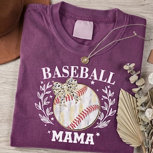 May include: A plum-colored crew neck t-shirt featuring a graphic of a baseball with a leopard print bow, surrounded by white laurel branches and the text BASEBALL MAMA in white capital letters. A thin gold-toned chain necklace rests on the shirt.