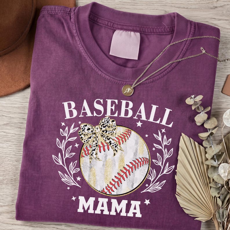 May include: A plum-colored crew neck t-shirt featuring a graphic of a baseball with a leopard print bow, surrounded by white laurel branches and the text BASEBALL MAMA in white capital letters. A thin gold-toned chain necklace rests on the shirt.