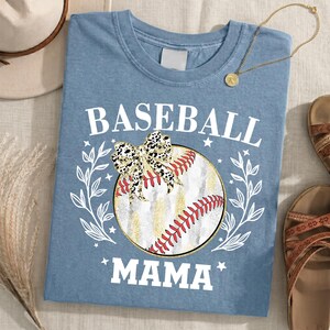May include: A dusty blue t-shirt featuring a graphic of a baseball with red stitching, a leopard print bow, and white leafy branches. The text BASEBALL MAMA is printed in white block letters above and below the central baseball illustration.