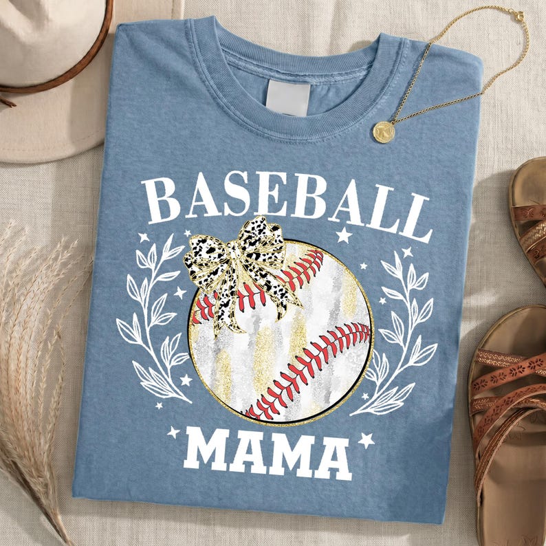 May include: A dusty blue t-shirt featuring a graphic of a baseball with red stitching, a leopard print bow, and white leafy branches. The text BASEBALL MAMA is printed in white block letters above and below the central baseball illustration.