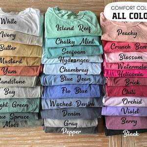 May include: A collection of folded t-shirts in various colors arranged in three vertical stacks. Each shirt features black text identifying its specific color name, such as White, Ivory, Butter, Mustard, Yam, Sandstone, Bay, Light Green, Blue Spruce, Moss, Island Reef, Chalky Mint, Seafoam, Hydrangea, Chambray, Blue Jean, Flo Blue, Washed Denim, Denim, Gray, Pepper, Peachy, Crunch Berry, Blossom, Watermelon, Heliconia, Brick, Chili, Orchid, Violet, Berry, and Black.