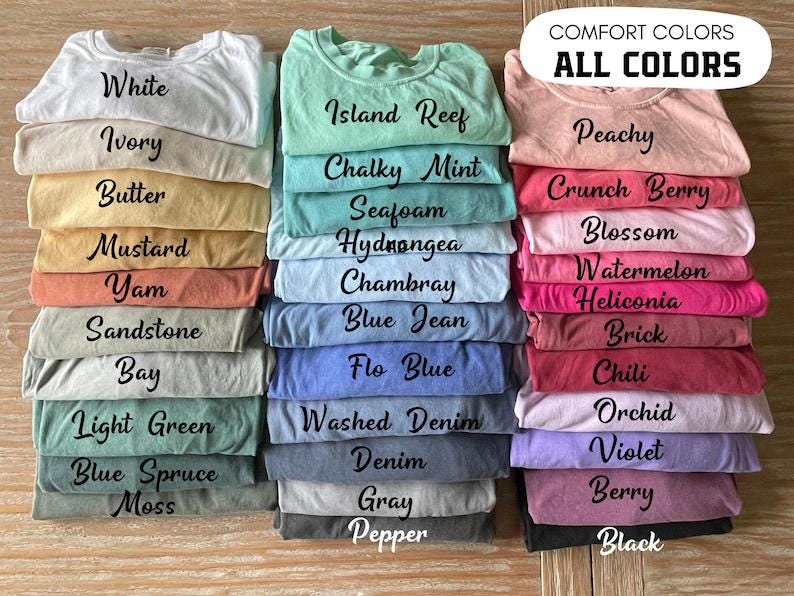 May include: A collection of folded t-shirts in various colors arranged in three vertical stacks. Each shirt features black text identifying its specific color name, such as White, Ivory, Butter, Mustard, Yam, Sandstone, Bay, Light Green, Blue Spruce, Moss, Island Reef, Chalky Mint, Seafoam, Hydrangea, Chambray, Blue Jean, Flo Blue, Washed Denim, Denim, Gray, Pepper, Peachy, Crunch Berry, Blossom, Watermelon, Heliconia, Brick, Chili, Orchid, Violet, Berry, and Black.