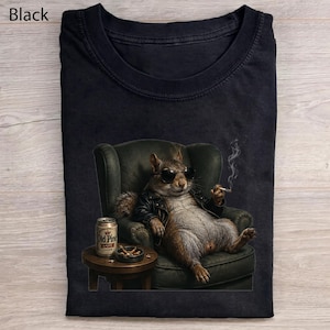 Może przedstawiać: Czarny t-shirt z grafiką wiewiórki w czarnej skórzanej kurtce i okularach przeciwsłonecznych, siedzącej w ciemnozielonym fotelu. Wiewiórka trzyma dymiącego papierosa, a na stoliku obok stoi puszka piwa z napisem Old Pine Lager.