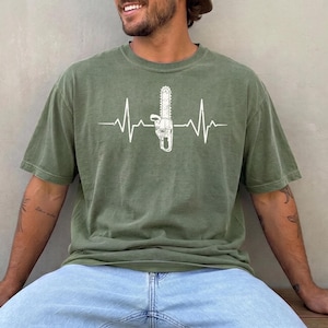 Op de afbeelding: Een olijfgroen T-shirt met korte mouwen en een witte grafische print van een hartslaglijn, of ECG, met een kettingzaag geïntegreerd in het midden van de hartslag. Het T-shirt heeft een ronde hals en een relaxte, casual pasvorm.