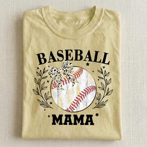 May include: A pale yellow t-shirt featuring a graphic of a baseball with red stitching, a leopard print bow, and black leaf accents. The text BASEBALL MAMA is printed in black capital letters above and below the central baseball illustration.