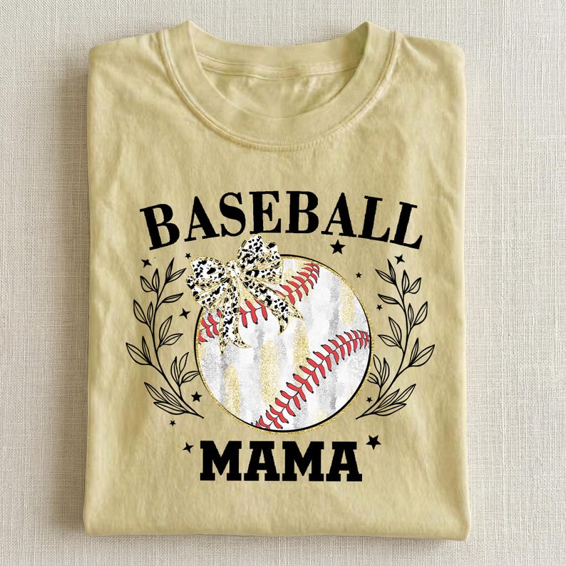 May include: A pale yellow t-shirt featuring a graphic of a baseball with red stitching, a leopard print bow, and black leaf accents. The text BASEBALL MAMA is printed in black capital letters above and below the central baseball illustration.