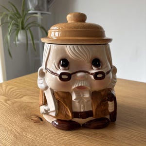 May include: A ceramic biscuit jar shaped like an elderly person wearing a tan hat, brown jacket, and glasses. The figure has white hair, a white moustache, and a bow tie. The jar features a removable lid and is painted in shades of tan, brown, and white.
