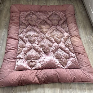 May include: A rectangular mauve-coloured quilted comforter featuring a smooth border and a central panel decorated with a repeating pattern of raised, textured leaf-shaped motifs. The fabric has a subtle sheen and a soft, padded construction.