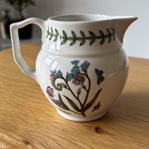 May include: A white ceramic cream jug featuring a botanical illustration of blue and pink flowers, green leaves, and small butterflies. The rim is decorated with a green leaf garland pattern, and the jug has a single angular handle.