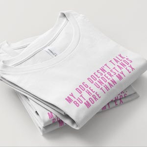 May include: A stack of white t-shirts with the text "MY DOG DOESN'T TALK. BUT HE UNDERSTANDS MORE THAN MY EX" printed in pink. The shirts are folded neatly, showcasing the crew neck design.