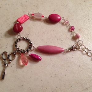 May include: A pink and silver bracelet with a charm of a pair of scissors. The bracelet has a variety of pink beads, including round, oval, and rectangular shapes.