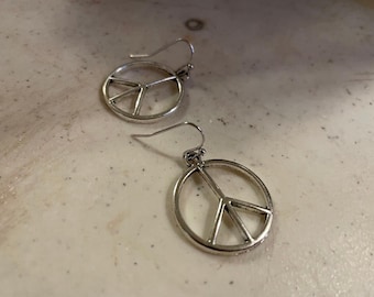 Może przedstawiać: Para srebrnych kolczyków w kształcie symbolu pokoju. Kolczyki są proste i eleganckie, z delikatnym wzorem symbolu pokoju.