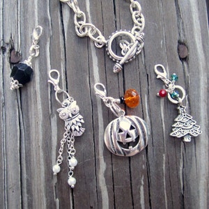 May include: A collection of silver charms and a bracelet with a toggle clasp. Charms include a black faceted bead, an owl with dangling chains and pearls, a jack-o'-lantern, and a Christmas tree with red and green accents. The charms are on a wooden surface.