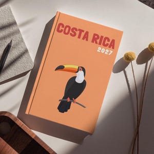 Peut inclure: Un journal orange avec les mots "COSTA RICA 2027" et une illustration de toucan. Un crayon noir et un carnet beige sont à côté du journal. Un plateau en bois et des fleurs jaunes sont également dans l'image.
