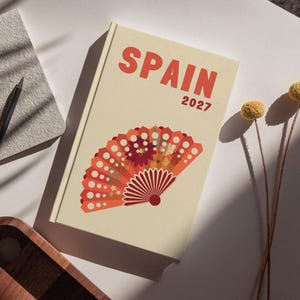 Peut inclure: Un carnet crème avec le mot "SPAIN" et l'année "2027" imprimés en rouge. La couverture présente un éventail décoratif orange et rouge. Un carnet gris, un crayon noir et un plateau en bois sont également visibles.