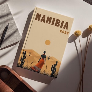 Puede incluir: Un diario beige titulado "NAMIBIA 2026" con una ilustración de paisaje desértico. La portada representa a una persona con un vestido naranja, cactus y un sol poniente. También se ven un cuaderno gris, un lápiz negro y una bandeja de madera.