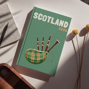 Peut inclure: Un livre vert avec le mot "SCOTLAND" en blanc et l'année "2026" en orange. La couverture présente un dessin de cornemuse avec un motif écossais. Un crayon, un carnet et un plateau en bois sont également visibles.