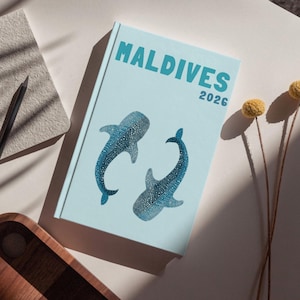 May include: A light blue notebook with the word "MALDIVES" and the year "2026" printed on the cover. Two blue whale shark illustrations are also on the cover. A gray notebook, a black pencil, and a wooden tray are also in the image.