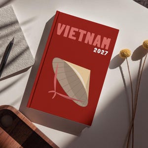 Peut inclure: Un livre rouge intitulé "VIETNAM 2027" avec une illustration d'un chapeau conique vietnamien. Il est posé sur une surface blanche, à côté d'un carnet gris, d'un crayon noir, d'un plateau en bois et de fleurs jaunes séchées.