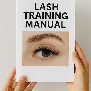 May include: A white book titled "LASH TRAINING MANUAL" is held by two hands. The book features a close-up image of an eye with long, dark eyelashes and a well-defined eyebrow. The eye is a light brown color.