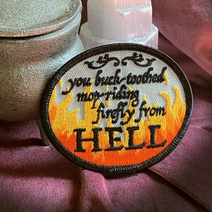 May include: A black and white embroidered patch with orange flames. The text on the patch reads "You buck-toothed mop-riding firefly from HELL".