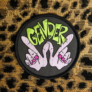 May include: A black and green embroidered patch with the word "GENDER" in green and two pink hands making the peace sign.