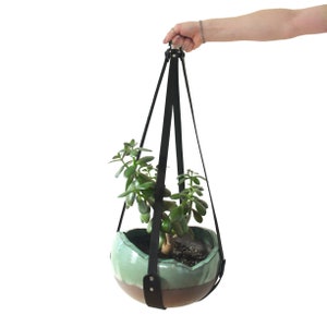 Puede incluir: Un macetero colgante de cuero negro con una maceta de cerámica verde y marrón y una pequeña planta verde en su interior.