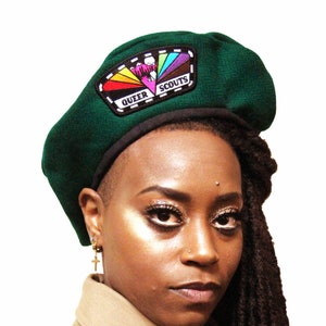 Peut inclure: Un béret vert foncé avec une bordure noire et un écusson avec l'inscription "QUEER SCOUTS" et un motif arc-en-ciel. Le béret est fait d'un tissu texturé.