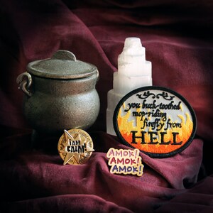 May include: An assortment of mystical items on a burgundy cloth. Includes a small, dark metal cauldron, a white crystal tower, a patch with the text "you buck-toothed mop-riding firefly from HELL", and two pins with text.