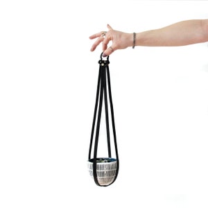 Puede incluir: Un colgador de plantas de cuero negro con una maceta de cerámica blanca. La maceta tiene un patrón geométrico y está colgada de las correas de cuero.