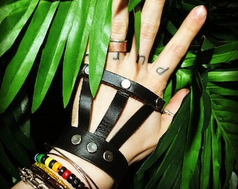 May include: A black leather fingerless glove with a strap around the wrist. The glove has a criss-cross design and is adorned with silver metal accents. The glove is worn on a hand with multiple bracelets, including a colorful beaded bracelet.