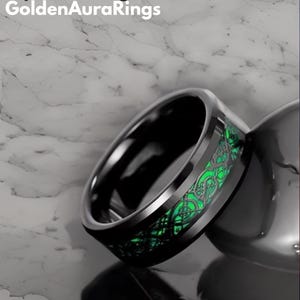 Puede incluir: Un anillo negro con un diseño verde de inspiración celta. El anillo tiene un acabado pulido y está sobre un fondo de mármol. El texto "Golden Aura Rings" está en la esquina superior izquierda.