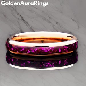Puede incluir: Un anillo de oro rosa con una banda con incrustaciones moteadas de color púrpura intenso y rosa. El anillo se refleja en la superficie inferior. El texto "Golden Aura Rings" está en la esquina superior izquierda.