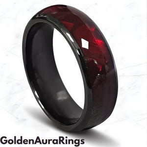Op de afbeelding: Een zwarte ring met een gefacetteerd, dieprood middenstuk. De ring heeft een glanzende afwerking en een gladde, afgeronde band. De tekst "Golden Aura Rings" staat onderaan de afbeelding.