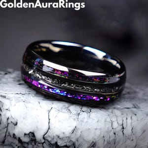 Puede incluir: Un anillo negro con un acabado pulido, con una banda de piedras moradas y azules iridiscentes. El anillo tiene una banda plateada con un diseño intrincado. El nombre de la marca "Golden Aura Rings" es visible en la parte superior.