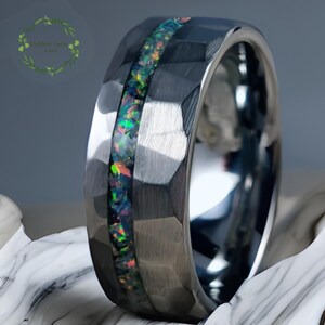 May include: A silver ring with a hammered texture and a central inlay of iridescent opal. The opal displays a vibrant array of colors, including green, blue, and red. The ring has a polished, reflective finish.
