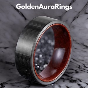以下が含まれることがあります： 黒いカーボンファイバーの外側と赤褐色の木材の内側を備えたリングのクローズアップ。 リングは、外側と内側の間に銀色のバンドがあります。 テキスト「GoldenAuraRings」が上部にあります。