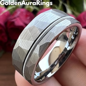 Puede incluir: Un anillo plateado con un diseño geométrico texturizado y una banda central de metal enrollado. El interior del anillo está pulido. El texto "GoldenAuraRings" es visible en la parte superior.