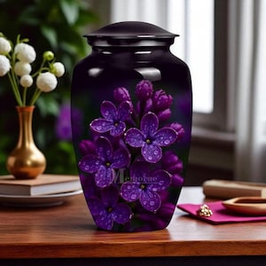 Op de afbeelding: Een donkerpaarse urn met een glanzende afwerking, met een levendige afbeelding van paarse seringenbloemen met waterdruppels. Het woord "Memorie" staat op de urn gedrukt. De urn is een gedenkitem.