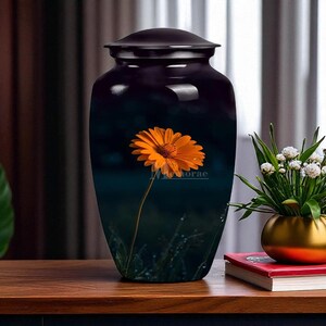 Op de afbeelding: Een donkere, glanzende urn met een deksel, met een oranje bloemdesign. De urn staat tegen een donkere achtergrond, met een bloemen- en grasillustratie. De urn staat op een houten oppervlak.