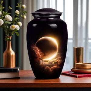 Puede incluir: Una urna funeraria negra con tapa, con un diseño celestial de un planeta y nubes con una luz brillante. La palabra "Memorae" está impresa en la urna. La urna está sobre una superficie de madera.