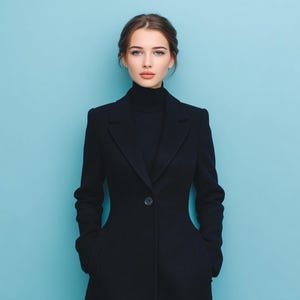 May include: A woman wearing a black coat and turtleneck against a light blue background. The coat has a single button closure and pockets. The woman has brown hair and is looking at the camera.