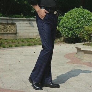 May include: Navy blue pinstripe trousers with a slight flare at the bottom. The trousers have vertical white stripes and are paired with black dress shoes. The person is wearing a black belt and a black shirt.