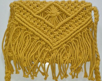 May include: Mustard yellow macrame shoulder bag with a geometric diamond pattern on the front. The bag features a braided strap and fringe along the bottom edge. This handmade bag is a stylish accessory.