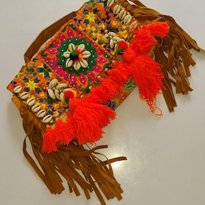Puede incluir: Un bolso de mano bordado y colorido con un vibrante estampado floral en naranja, amarillo y verde. La bolsa está adornada con conchas marinas y borlas de color naranja brillante. Flecos de ante marrón cuelgan de la parte inferior y una correa marrón es visible.