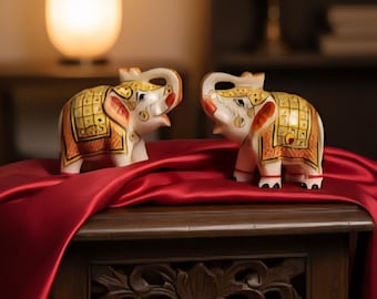 Puede incluir: Dos figuras decorativas de elefantes. Los elefantes son blancos con detalles dorados y naranjas, y están colocados sobre una mesa de madera oscura cubierta con una tela de satén roja. Una luz suave ilumina la escena desde atrás.