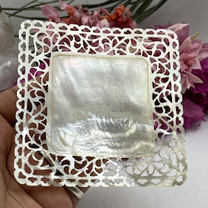 May include: A square tray featuring a smooth, iridescent mother-of-pearl centre panel framed by an intricate, white, laser-cut filigree border. The material displays a natural, pearlescent sheen with subtle variations in texture and light reflection.
