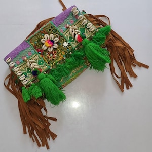 Peut inclure: Une pochette de style bohème colorée avec un design patchwork. Le sac présente des tissus verts, violets et dorés, ornés de coquillages, de perles et de pompons. Des franges en daim marron pendent sur les côtés, ajoutant un élément texturé à l'accessoire.